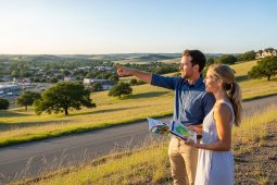 Um homem e uma mulher jovens estão de pé no topo de uma colina gramada durante o pôr do sol, observando uma vista panorâmica de um vale com pequenas construções e árvores. O homem aponta para o horizonte enquanto a mulher segura um folheto e um tablet que exibe um mapa. Ambos sorriem, transmitindo uma sensação de otimismo e planejamento para o futuro em um ambiente natural e tranquilo